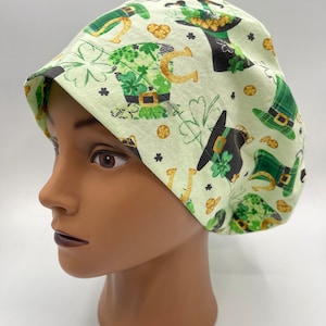May include: A green and gold scrub hat with a pattern of shamrocks, horseshoes, and leprechaun hats. The hat is on a mannequin head.