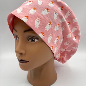May include: Pink scrub cap with a repeating pattern of white ghosts wearing orange cowboy hats. The cap is made of fabric and is designed to cover the head and hair.