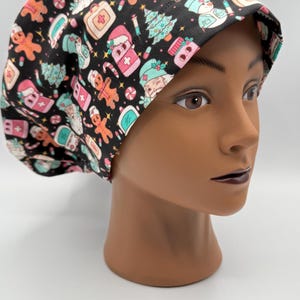 May include: A black scrub cap featuring a festive Christmas pattern. The design includes gingerbread men, Christmas trees, and medical-themed elements in pink, teal, and white. The cap is worn by a mannequin head.