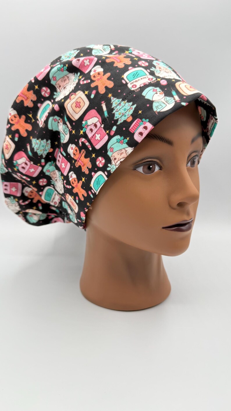 May include: A black scrub cap featuring a festive Christmas pattern. The design includes gingerbread men, Christmas trees, and medical-themed elements in pink, teal, and white. The cap is worn by a mannequin head.