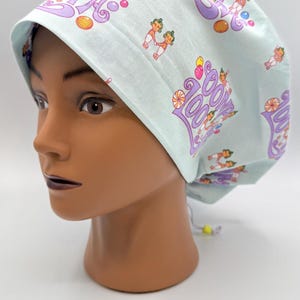 May include: A light blue scrub cap with a repeating pattern of cartoon figures, candies, and the words "Spa Zoom". The cap is on a mannequin head. The cap has a drawstring closure.
