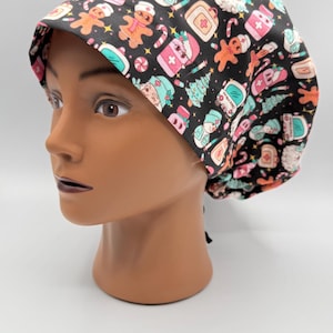 May include: A black scrub cap with a festive Christmas pattern. The cap features gingerbread men, Christmas trees, and medical-themed icons in pink, teal, and white. The cap is displayed on a mannequin head.