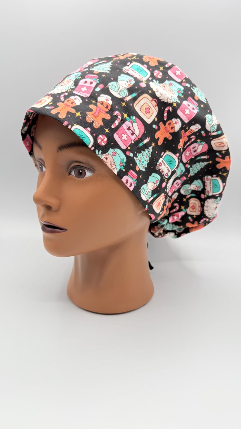 May include: A black scrub cap with a festive Christmas pattern. The cap features gingerbread men, Christmas trees, and medical-themed icons in pink, teal, and white. The cap is displayed on a mannequin head.