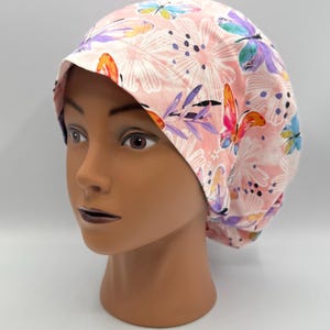May include: A pink surgical scrub cap with a floral and butterfly pattern. The cap features a repeating design of white flowers, purple leaves, and colorful butterflies in shades of orange, blue, and pink. The cap is displayed on a mannequin head.