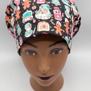 May include: A black scrub cap with a colorful Christmas-themed pattern. The pattern includes gingerbread men, Santa Claus, Christmas trees, and medical symbols. The cap is designed to be worn in a medical setting.