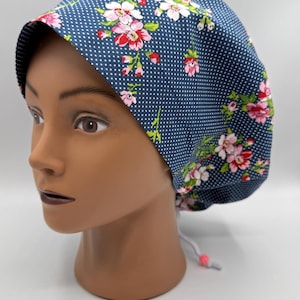 May include: A navy blue scrub cap with a white polka dot pattern and floral accents in pink and white. The cap is tied at the back with a white drawstring and a pink bead.