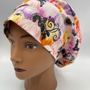 Puede incluir: Un gorro de quirófano blanco con un estampado floral colorido que presenta calabazas, murciélagos y remolinos. El estampado es en tonos naranja, rosa, morado y negro.