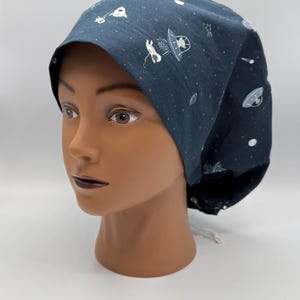 Puede incluir: Un gorro quirúrgico azul oscuro con un patrón repetido de ilustraciones blancas con temática espacial, que incluyen planetas, estrellas y ovnis. El gorro se muestra en una cabeza de maniquí.