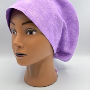 May include: A light purple scrub cap with a heart and swirl pattern. The cap is on a mannequin head. The cap is made of fabric and has a brim. The background is white.