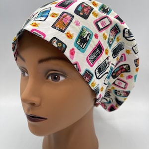 May include: A white surgical scrub cap with a repeating pattern of smartphones, cats, and paw prints in pink, black, and teal. The cap is made of a soft fabric and is designed to cover the hair during medical procedures.
