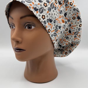 May include: A floral patterned scrub cap in a light gray color. The cap features a repeating pattern of black and orange flowers and leaves. The cap is designed to be worn in a medical or professional setting.