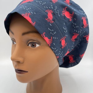 May include: A navy blue scrub cap featuring a repeating pattern of red crabs and white dashes. The cap is made of fabric and has a rounded shape, designed to cover the head. The cap is on a mannequin head.