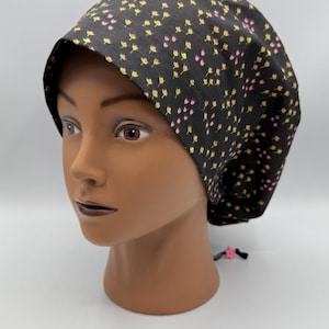 May include: A black surgical scrub cap with a pattern of small pink and yellow flowers. The cap is tied at the back with a black string and a pink flower accent. The cap is on a mannequin head.