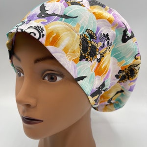 Puede incluir: Un gorro de quirófano blanco con un estampado colorido de calabazas, murciélagos y girasoles. El estampado es en tonos naranja, morado, verde y negro.