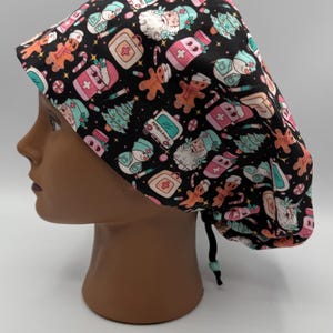 May include: A black scrub cap featuring a festive Christmas pattern. The design includes gingerbread men, snowmen, Christmas trees, and medical-themed icons in pink, teal, and white. The cap is tied at the back with a black drawstring.