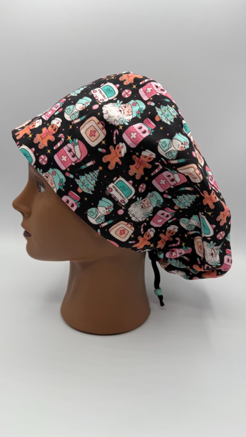 May include: A black scrub cap featuring a festive Christmas pattern. The design includes gingerbread men, snowmen, Christmas trees, and medical-themed icons in pink, teal, and white. The cap is tied at the back with a black drawstring.