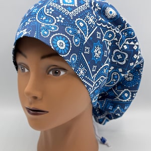 May include: A blue scrub cap featuring a white and dark blue paisley and floral pattern. The cap has a rounded crown and a gathered back section with a white drawstring cord and a small blue plastic toggle for an adjustable fit around the head.