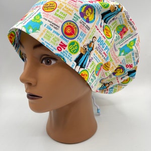 May include: A white scrub hat with a colorful print featuring quotes and images from the movie "The Wizard of Oz". The print includes phrases like "A real heart", "I like your shoes", and "No place like home".
