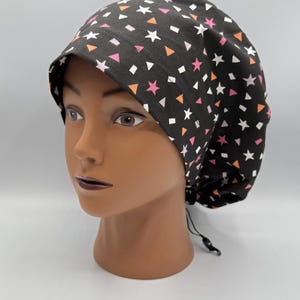 Puede incluir: Un gorro quirúrgico negro con un estampado de estrellas blancas, estrellas rosas, triángulos naranjas y cuadrados blancos. El gorro está sobre una cabeza de maniquí. Tiene un cierre de cordón.