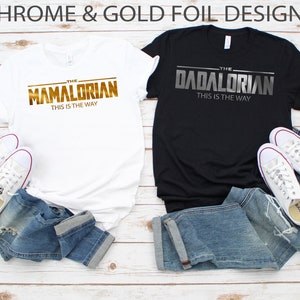 May include: Two t-shirts with a Star Wars theme. One white t-shirt with gold foil lettering that says "The Mamalorian This is the Way". One black t-shirt with silver foil lettering that says "The Dadalorian This is the Way".