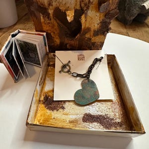 May include: A heart-shaped necklace with a turquoise patina finish, on a chain with a toggle clasp. The necklace is displayed in a small, white, metal box with a rusty brown finish. The box has a white card inside with the text "Carnival of Rust" printed on it.