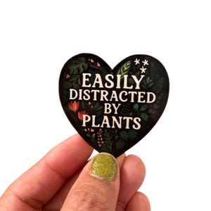 May include: A heart-shaped sticker with the text "EASILY DISTRACTED BY PLANTS" in white. The sticker has a black background with a botanical design of various plants and flowers in shades of green, red, and white.