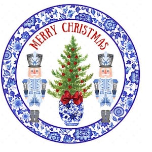 Puede incluir: Un plato blanco redondo con un diseño floral azul y blanco. El plato presenta dos figuras de cascanueces azules y blancas que flanquean un árbol de Navidad verde con bayas rojas. El texto "Merry Christmas" está escrito en letras rojas alrededor del árbol.