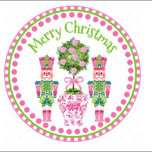 May include: A pink and green Christmas ornament featuring two nutcracker figurines flanking a pink and white floral patterned vase with a pink rose bush. The ornament has a white background with a pink and green polka dot border and the text "Merry Christmas" in green.