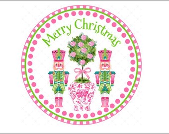 Puede incluir: Un adorno navideño rosa y verde con dos figuras de cascanueces a ambos lados de un jarrón de patrón floral rosa y blanco con un rosal rosa. El adorno tiene un fondo blanco con un borde de lunares rosa y verde y el texto "Merry Christmas" en verde.