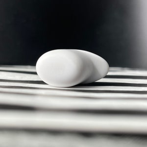 May include: A white, smooth, rounded earbud with a slightly pointed end. The earbud is lying on a black and white striped surface.