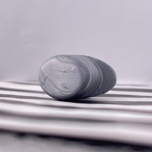 May include: A smooth, gray, oval-shaped stone with swirling patterns. The stone is resting on a white and gray striped surface.