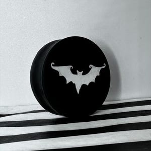 May include: Black round plug with a white bat design. The bat has a stylized wing shape. The plug is set against a black and white striped background, with a plain white wall in the background.