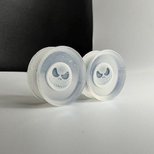 May include: A pair of translucent white ear plugs featuring a centered, raised white circular emblem with a black printed graphic of a grinning, wide-eyed skull face with stitched mouth details, designed for body modification jewelry.