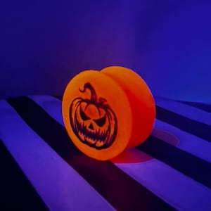 May include: A circular orange ear plug featuring a black graphic of a carved jack-o'-lantern pumpkin with a jagged, menacing face. The object glows with a bright orange hue against a dark blue background, highlighting the detailed pumpkin design.
