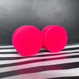 May include: Two vivid pink, circular objects with a subtle indentation, similar to a peach. They are positioned against a black and white striped background, with a dark grey backdrop.