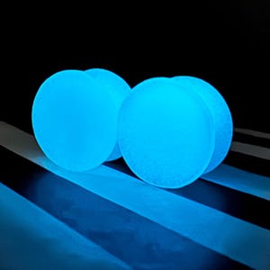Puede incluir: Cuatro esferas azules brillantes se agrupan, emitiendo una suave luz etérea. Descansan sobre una superficie con rayas blancas y negras, creando un contraste llamativo sobre un fondo oscuro. Las esferas parecen estar hechas de un material texturizado.