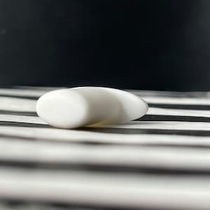 Può includere: Un oggetto bianco liscio a forma di goccia. L'oggetto ha un'estremità arrotondata e si assottiglia fino a un punto. È posto su uno sfondo a strisce bianche e nere, con uno sfondo nero.