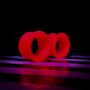 May include: Two bright red, cylindrical ear gauges are shown against a black background with purple and black stripes. The gauges are lit, creating a glowing effect. The image is a studio shot.