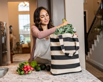 May include: A woman is putting groceries into a black and white striped canvas tote bag. The bag is sitting on a kitchen counter with fresh produce, including radishes, limes, and avocados.