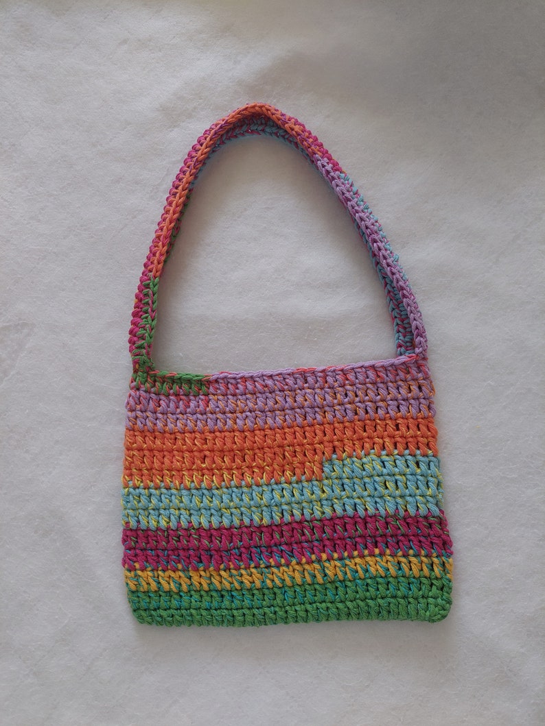 Op de afbeelding: Een veelkleurige gehaakte tote bag met een lange band. De tas is gemaakt van verschillende kleuren, waaronder groen, blauw, oranje, geel en roze.