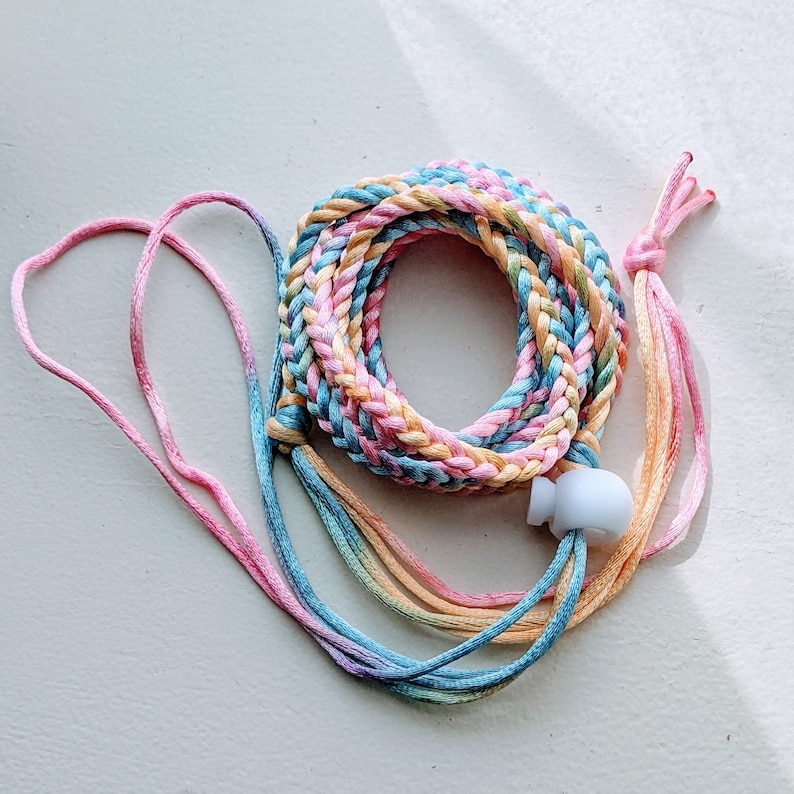 May include: A braided, multi-colored lanyard in pastel shades of pink, blue, yellow, and orange. The lanyard has a white plastic adjuster and pink and orange tassels at the ends. The braided section is approximately 10 inches in diameter.