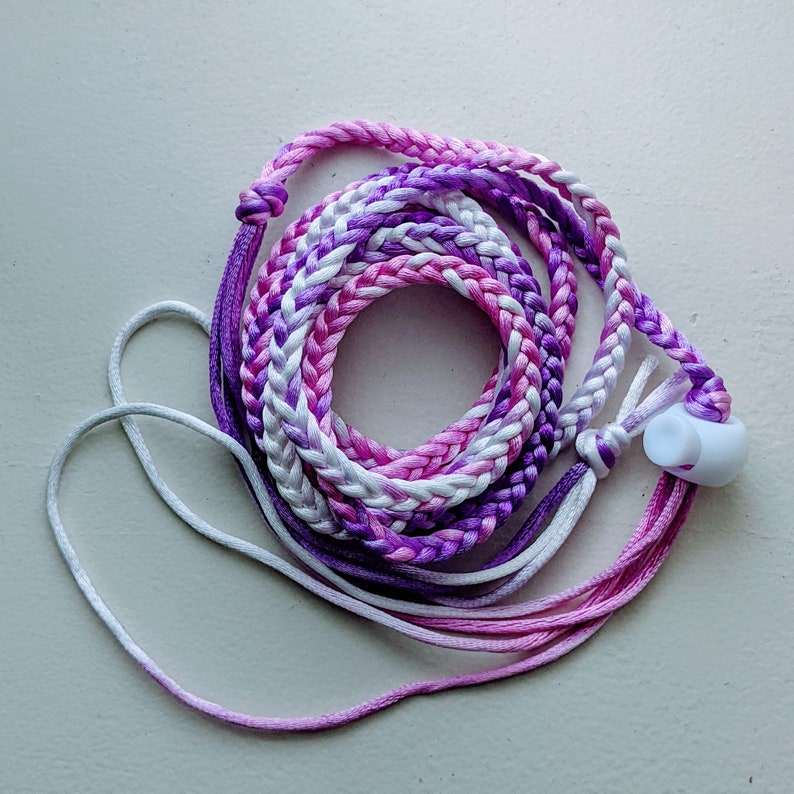 May include: Braided lanyard in shades of purple, pink, and white. The cord is coiled, with a white plastic clasp. The lanyard is designed to be worn around the neck.
