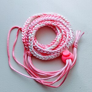 May include: A coiled pink and white braided cord with a pink plastic toggle. The cord is made of multiple strands of pink and white thread, creating a textured appearance. The toggle is a bright pink color.