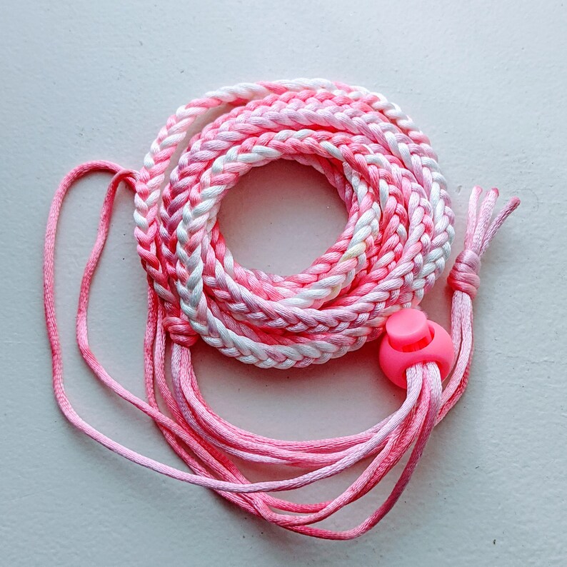 May include: A coiled pink and white braided cord with a pink plastic toggle. The cord is made of multiple strands of pink and white thread, creating a textured appearance. The toggle is a bright pink color.