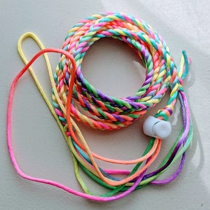 May include: A colorful braided lanyard with a white earbud. The lanyard is made of multiple strands of rainbow-colored cord, including pink, orange, yellow, green, blue, and purple. The earbud is white and has a smooth, rounded shape.