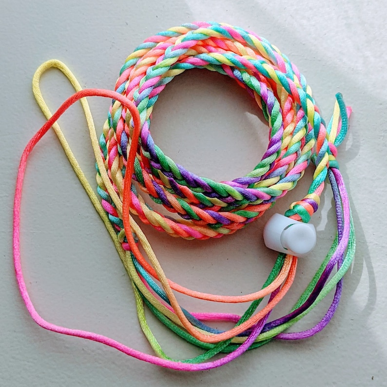 May include: A colorful braided lanyard with a white earbud. The lanyard is made of multiple strands of rainbow-colored cord, including pink, orange, yellow, green, blue, and purple. The earbud is white and has a smooth, rounded shape.