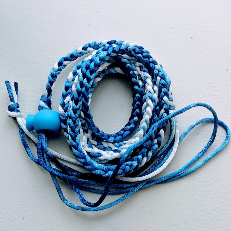 May include: Braided lanyard in shades of blue and white, coiled on a white surface. The lanyard features a blue bead and adjustable cords. The braided section is approximately 30 inches long.