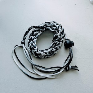 May include: Braided lanyard in shades of black, white, and gray. The cord is looped and coiled, with a black plastic adjuster and loose ends. The lanyard is designed to be worn around the neck.