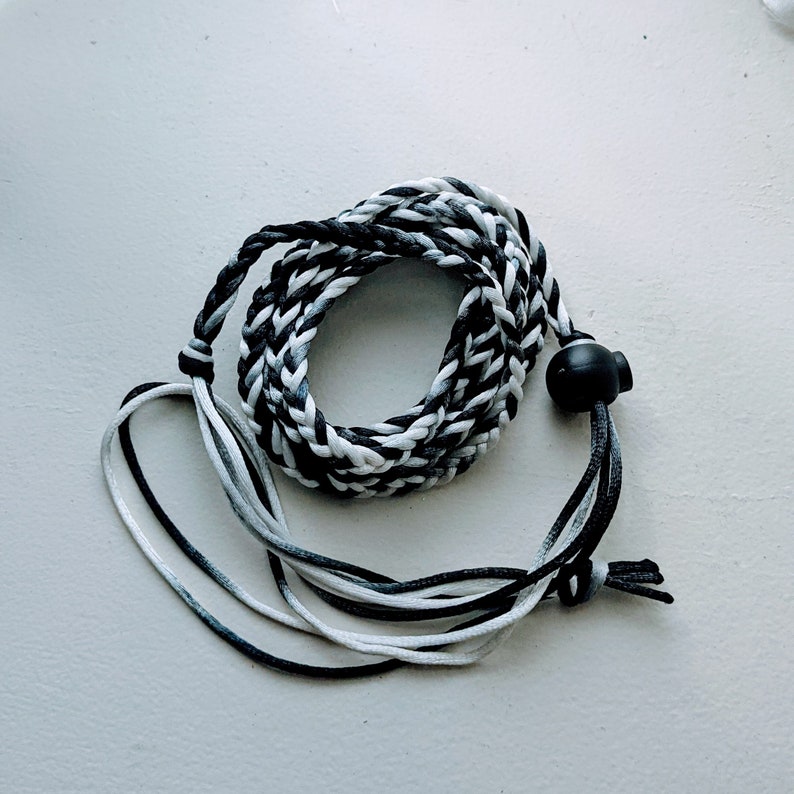 May include: Braided lanyard in shades of black, white, and gray. The cord is looped and coiled, with a black plastic adjuster and loose ends. The lanyard is designed to be worn around the neck.