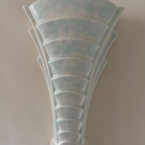 May include: A wall-mounted ceramic vase featuring a fan-shaped silhouette with tiered, horizontal ridges. The surface has a glossy, pale white finish with subtle, mottled light blue accents throughout the structure, tapering down to a narrow base.